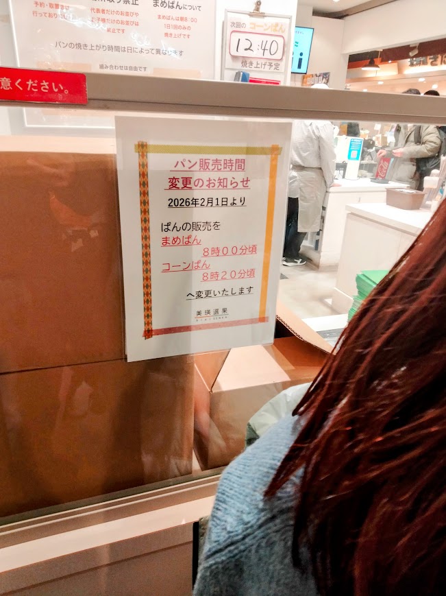 パン販売時間変更のお知らせの案内が店舗に貼られている。2,026年2月1日よりまめぱん8時ころ、コーンぱん8時20分頃へ変更しますと記載されている。
