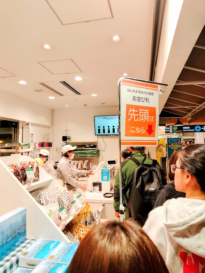 店舗レジ付近、先頭はこちらと書かれた列案内の看板と、列に並ぶ人々